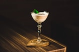 A cocktail glass filled with a creamy drink is elegantly placed on a wooden surface. The drink is garnished with a fresh green leaf. Soft lighting creates a warm and inviting atmosphere, highlighting the textured tabletop.