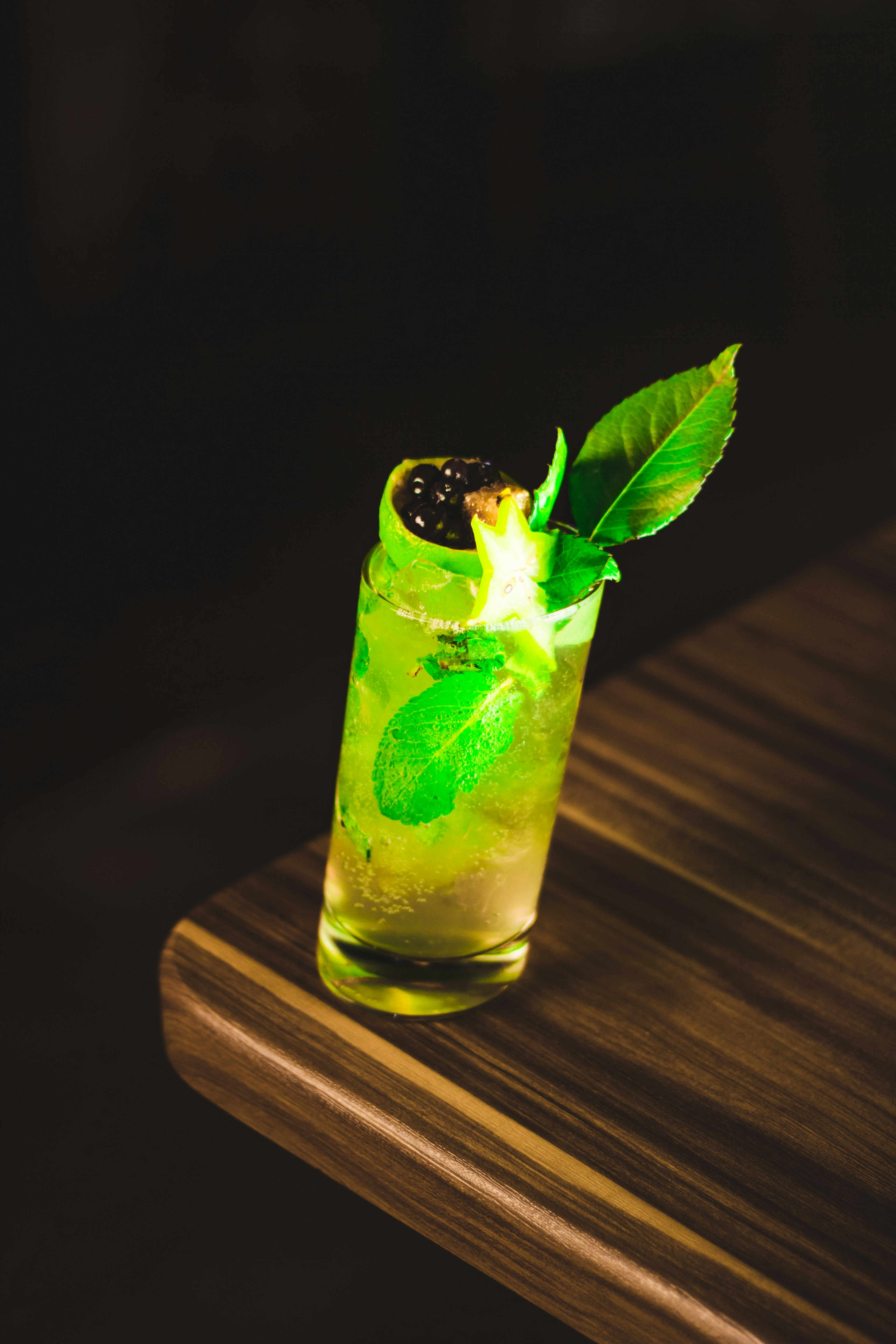 A cocktail glass sits on a wooden table, filled with a refreshing drink that includes lime wedges and mint leaves. The drink has a vibrant green hue and is garnished with a star fruit slice and blackberries.