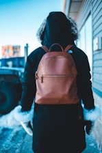 A person wearing a dark hooded parka and gloves with white fur trim is standing outdoors. They have a brown leather backpack on their back. The person is facing away, and the setting appears cold with snow on the ground. A building and a vehicle are partly visible in the background.