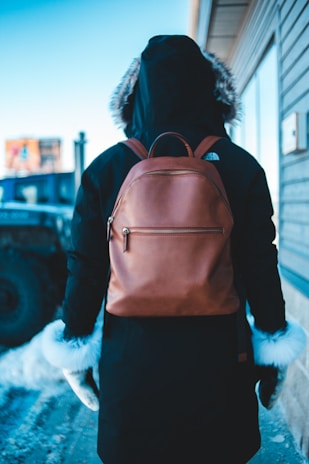 A person wearing a dark hooded parka and gloves with white fur trim is standing outdoors. They have a brown leather backpack on their back. The person is facing away, and the setting appears cold with snow on the ground. A building and a vehicle are partly visible in the background.