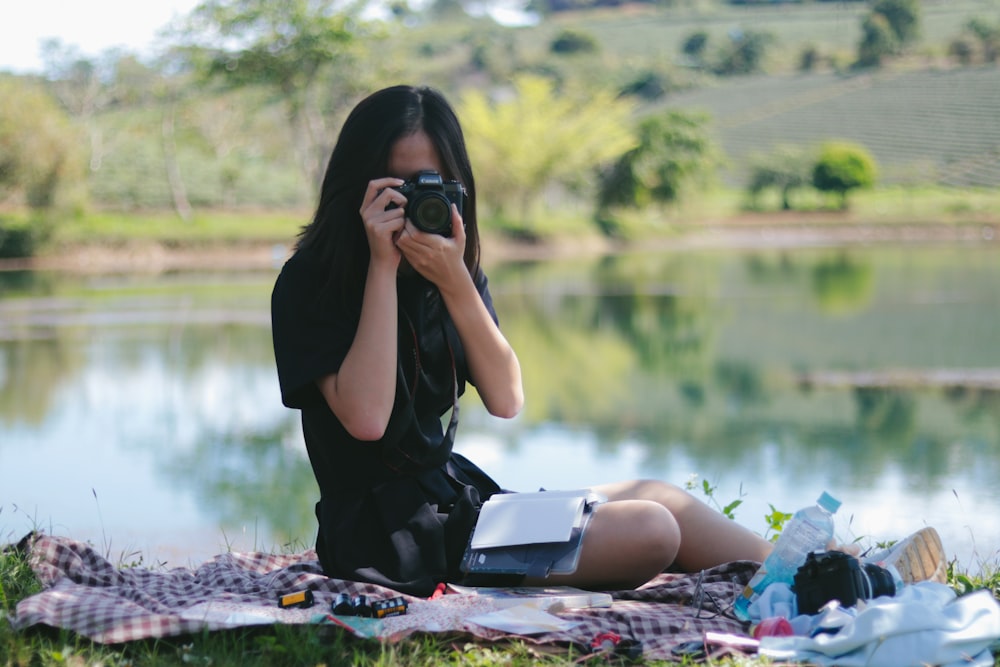 Frau mit Kamera, während sie auf einem Tuch auf Gras neben dem Gewässer sitzt