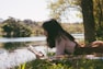 woman reading book and lying forward on sheet on grass beside body of water during day