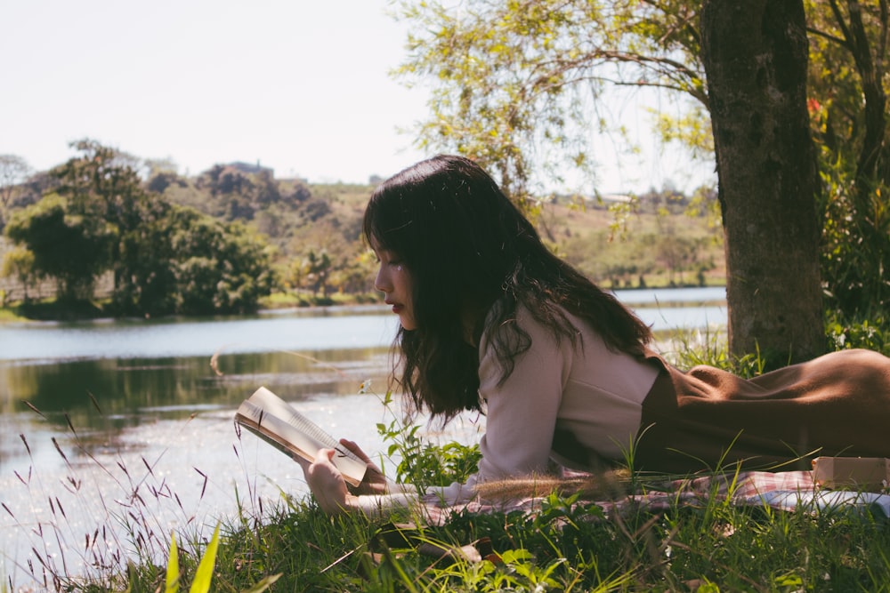 woman reading book and lying forward on sheet on grass beside body of water during day