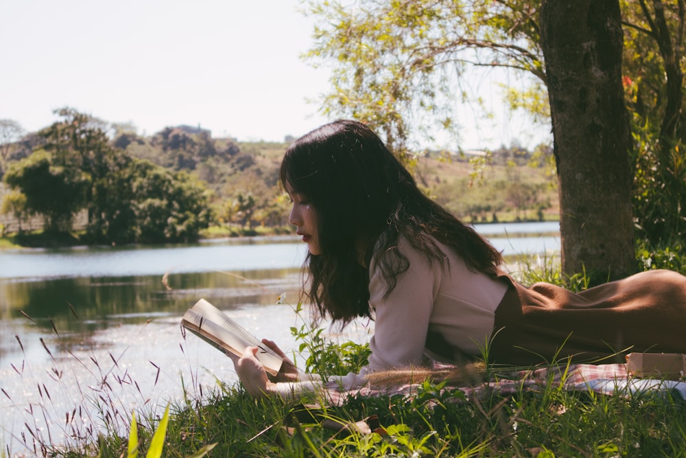 日中、水辺の芝生の上で本を読み、シートの上に前かがみに横たわる女性