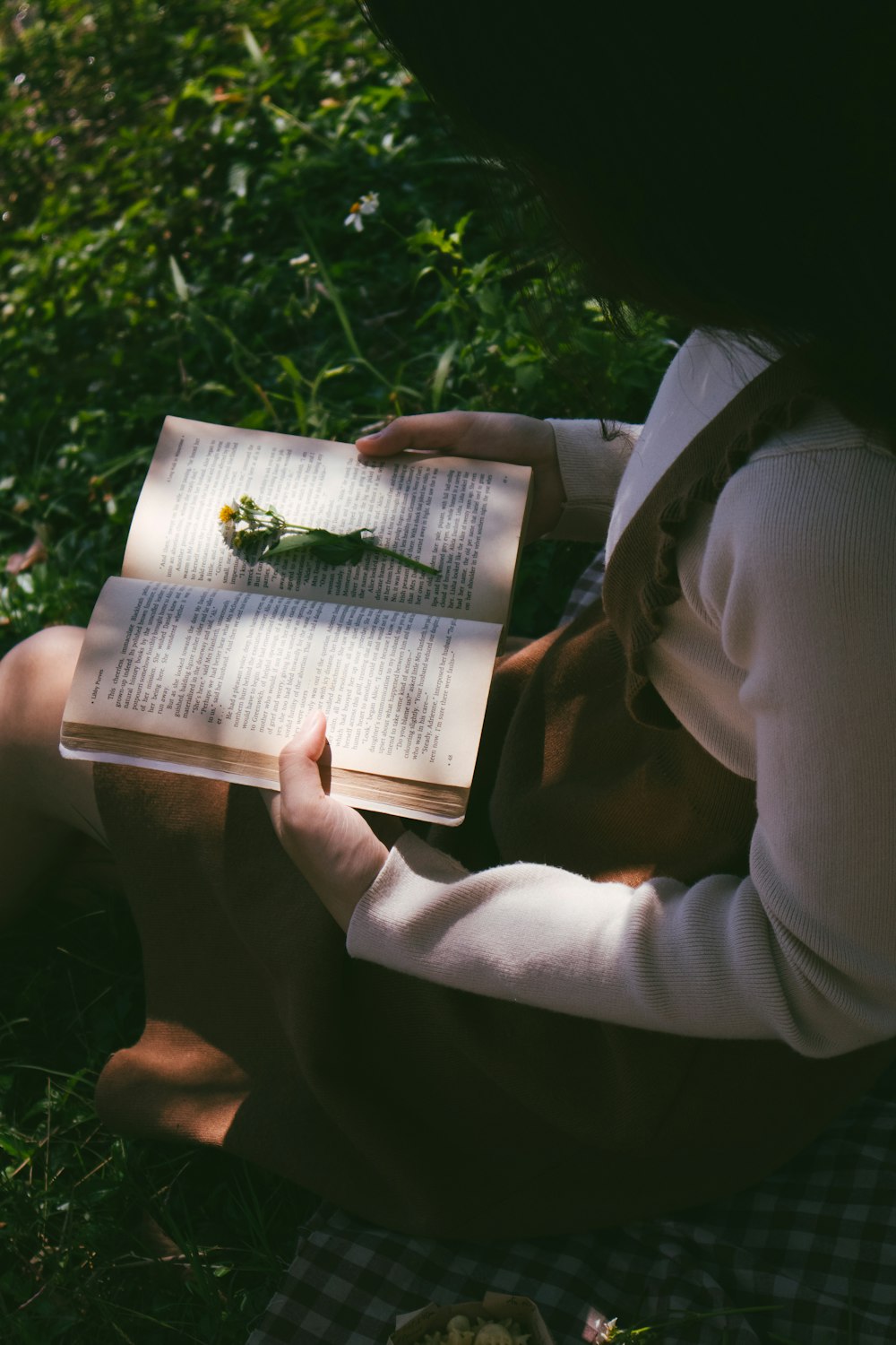 woman wearing white long-sleeved shirt sitting on green field while reading book and green flower on top of book