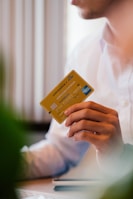 A person wearing a white shirt holds a yellow Air France KLM American Express card in their hand. The person is seated, possibly at a desk, with some blurred greenery in the foreground.