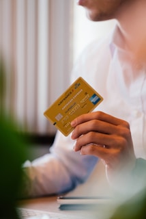 A person wearing a white shirt holds a yellow Air France KLM American Express card in their hand. The person is seated, possibly at a desk, with some blurred greenery in the foreground.