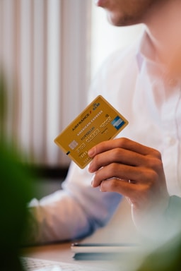 A person wearing a white shirt holds a yellow Air France KLM American Express card in their hand. The person is seated, possibly at a desk, with some blurred greenery in the foreground.