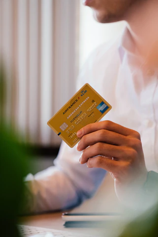 A person wearing a white shirt holds a yellow Air France KLM American Express card in their hand. The person is seated, possibly at a desk, with some blurred greenery in the foreground.