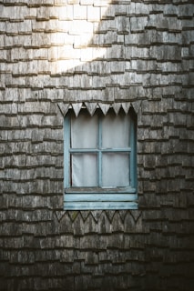 A wooden exterior wall is covered with weathered shingles, featuring a small window with a light blue frame in the center. Light filters onto the upper left corner, highlighting the texture of the shingles.