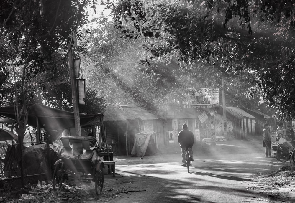 grayscale photography of man biking and other people walking on street near houses surrounded with trees