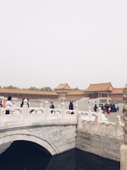 A historical architectural setting featuring a traditional Chinese palace with orange-tiled roofs and ornate white stone bridges. Several people are walking and standing along the bridge, taking in the surroundings.