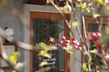 A café or smoothie shop entrance with a glass door displaying a hanging green wreath with red berries and white ornaments, partially obscured by a branch of a plant with round red fruits and green leaves in the foreground. The door features business hours, indicating the shop is open daily from 9:00 to 20:00.