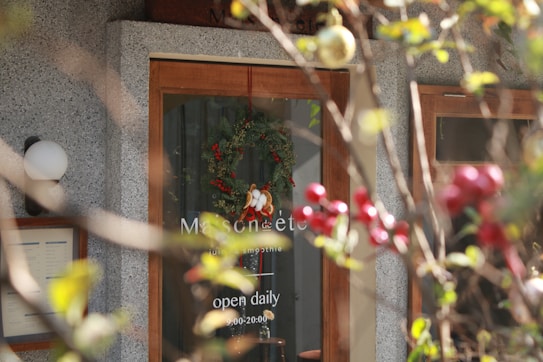 A caf&eacute; or smoothie shop entrance with a glass door displaying a hanging green wreath with red berries and white ornaments, partially obscured by a branch of a plant with round red fruits and green leaves in the foreground. The door features business hours, indicating the shop is open daily from 9:00 to 20:00.