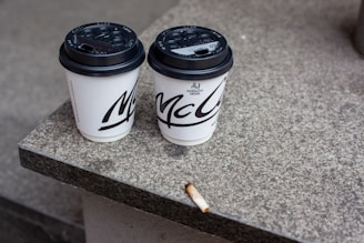 Two disposable coffee cups with black lids are placed side by side on a stone surface, with a cigarette butt lying nearby. The surface appears to be part of a step or ledge.