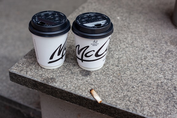 Two disposable coffee cups with black lids are placed side by side on a stone surface, with a cigarette butt lying nearby. The surface appears to be part of a step or ledge.