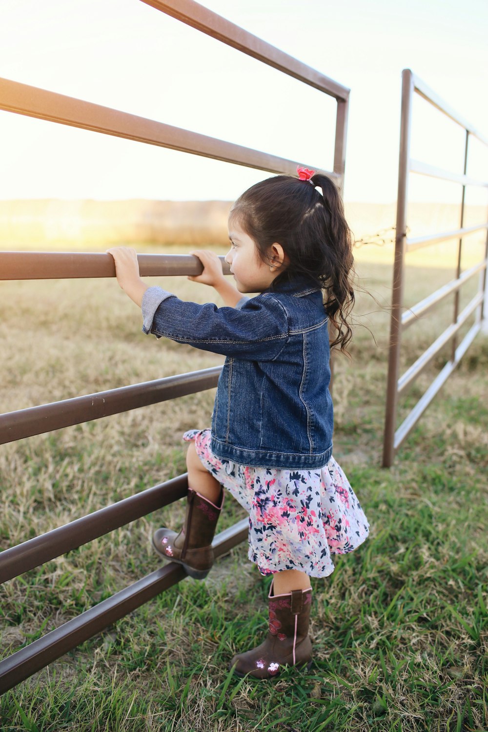 child wearing blue denim jacket standing and holding on wooden fence during daytime