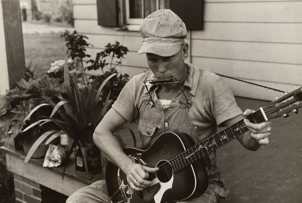 grayscale photography of man sitting while playing acoustic guitar