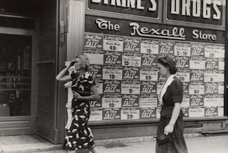 Two women walking past a storefront with extensive signage advertising various products on sale. The storefront has large advertisements with prices for items such as mouth wash, facial tissues, and tooth brushes. The store name, 'The Rexall Store,' is visible above the advertisements. One woman is wearing a dark dress with a pattern and the other a simpler dark dress.