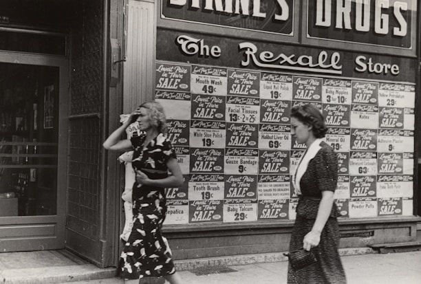 Two women walking past a storefront with extensive signage advertising various products on sale. The storefront has large advertisements with prices for items such as mouth wash, facial tissues, and tooth brushes. The store name, 'The Rexall Store,' is visible above the advertisements. One woman is wearing a dark dress with a pattern and the other a simpler dark dress.