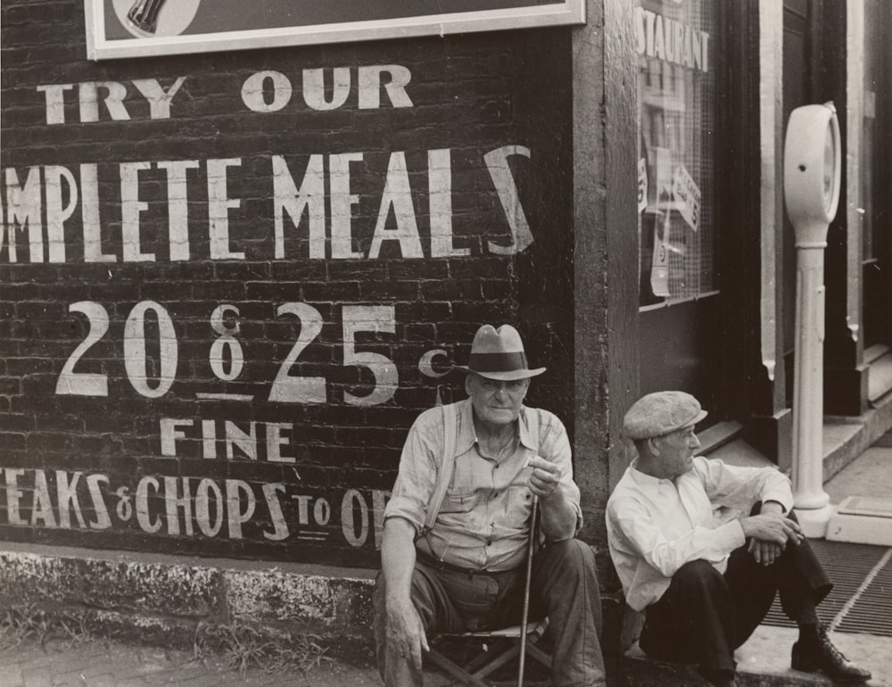 grayscale photography of two men sitting near concrete wall