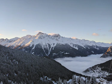 Snow-capped mountains with a cozy lodge nestled among pine trees under a clear blue sky.