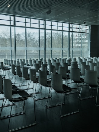 An empty conference room filled with rows of modern, minimalist chairs neatly arranged. Large ceiling-to-floor windows on one side let in natural light, casting soft shadows across the tiled floor. The view outside features a blurred urban landscape.
