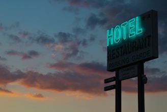 A neon sign for a hotel and restaurant is illuminated against a twilight sky filled with dark, scattered clouds. The sign displays the words 'HOTEL' and 'RESTAURANT' prominently, with smaller text below indicating parking options and room rates. The sky exhibits a gradient of blue and orange hues from the setting sun.