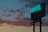 A neon sign for a hotel and restaurant is illuminated against a twilight sky filled with dark, scattered clouds. The sign displays the words 'HOTEL' and 'RESTAURANT' prominently, with smaller text below indicating parking options and room rates. The sky exhibits a gradient of blue and orange hues from the setting sun.