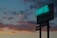 A neon sign for a hotel and restaurant is illuminated against a twilight sky filled with dark, scattered clouds. The sign displays the words 'HOTEL' and 'RESTAURANT' prominently, with smaller text below indicating parking options and room rates. The sky exhibits a gradient of blue and orange hues from the setting sun.