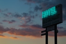 A neon sign for a hotel and restaurant is illuminated against a twilight sky filled with dark, scattered clouds. The sign displays the words 'HOTEL' and 'RESTAURANT' prominently, with smaller text below indicating parking options and room rates. The sky exhibits a gradient of blue and orange hues from the setting sun.