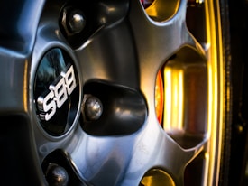 Close-up view of a car wheel with a metallic finish featuring a BBS logo in the center. The image highlights the detailed design of the wheel spokes and the reflective surface, with hints of surrounding light casting a yellowish glow.