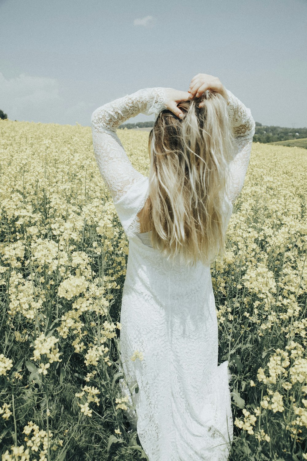 woman wearing white long-sleeved dress facing back while standing near flower field and putting both hands on her head under white and blue sky