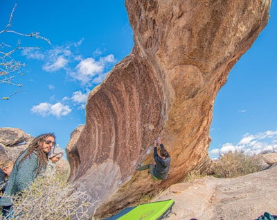 A person is bouldering on a large, overhanging rock formation in a sunny outdoor setting. Another person is observing while standing nearby, and there is climbing gear on the ground including a crash pad. The sky is clear with a few scattered clouds and the area is surrounded by rocks and sparse vegetation.