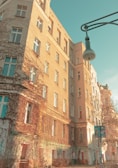 A tall, ivy-covered residential building with many windows is bathed in warm sunlight. The building features a vintage street lamp and a tree casting shadows. There are signs of urban life, such as graffiti and a bicycle parked at the base of the building.