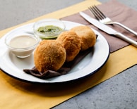 Three breaded and fried items are placed on a white plate with a dark napkin. Two small glass bowls accompany the dish, one filled with a green sauce and the other with a creamy white sauce. A knife and fork are set to the right of the plate on a brown napkin, resting on a textured gray surface.