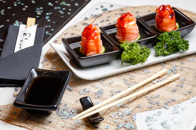 Three black square dishes are arranged on a white tray, each containing a piece of sushi topped with a bright red sauce, placed on cucumber slices. Garnishes of fresh green parsley accompany the sushi. A small rectangular dish is filled with soy sauce, placed beside a pair of wooden chopsticks resting on a textured mat. Black napkins with chopsticks wrapped in paper rest nearby. The table is decorated with scattered sparkling sequins.