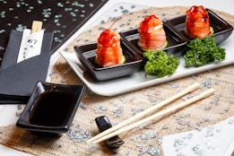 Three black square dishes are arranged on a white tray, each containing a piece of sushi topped with a bright red sauce, placed on cucumber slices. Garnishes of fresh green parsley accompany the sushi. A small rectangular dish is filled with soy sauce, placed beside a pair of wooden chopsticks resting on a textured mat. Black napkins with chopsticks wrapped in paper rest nearby. The table is decorated with scattered sparkling sequins.