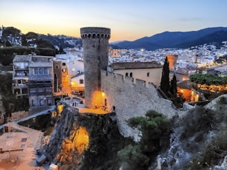 A picturesque medieval town at dusk, featuring a towering stone castle wall and turrets overlooking a bustling village below. The area is illuminated with warm, ambient lighting, highlighting the ancient architecture and cobbled streets. Surrounding mountains are visible, adding depth to the scene.