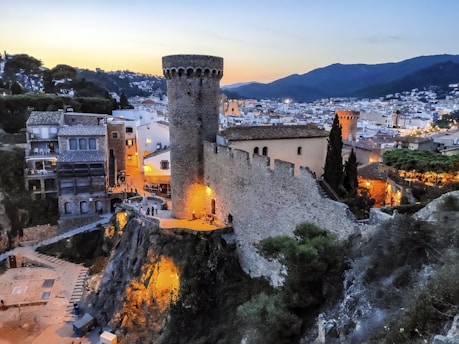 A picturesque medieval town at dusk, featuring a towering stone castle wall and turrets overlooking a bustling village below. The area is illuminated with warm, ambient lighting, highlighting the ancient architecture and cobbled streets. Surrounding mountains are visible, adding depth to the scene.