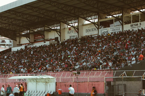 A crowded stadium with spectators filling the stands, observing an event. The stadium is covered and has multiple sections labeled with various advertisements visible on the walls. In front of the stands, a few people are gathered near an area with chairs and a white canopy.