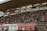 A crowded stadium with spectators filling the stands, observing an event. The stadium is covered and has multiple sections labeled with various advertisements visible on the walls. In front of the stands, a few people are gathered near an area with chairs and a white canopy.