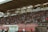 A crowded stadium with spectators filling the stands, observing an event. The stadium is covered and has multiple sections labeled with various advertisements visible on the walls. In front of the stands, a few people are gathered near an area with chairs and a white canopy.