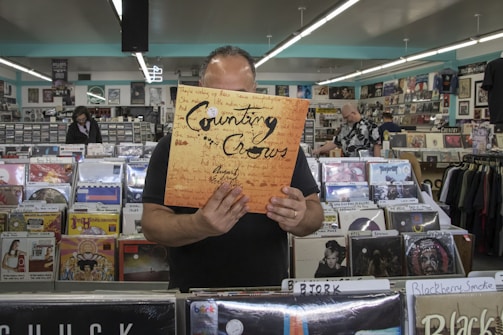 A person holds a Counting Crows vinyl record in a record store. The store is filled with various vinyl records displayed in bins and on shelves. Other customers are browsing in the background, surrounded by vibrant posters and music-related decor on the walls.