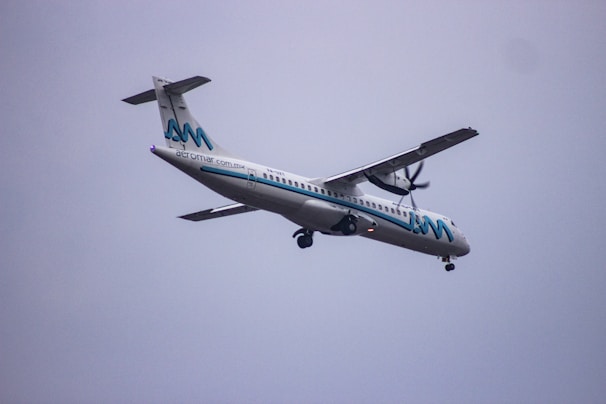 A propeller-driven passenger airplane, branded with the name Aeromar, is flying through a cloudy sky. The aircraft is white with blue and black accents, featuring a logo on the tail and the company name along the fuselage.