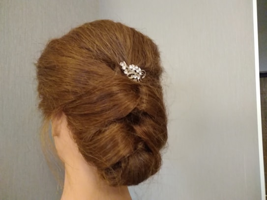 A neatly styled updo with brown hair, featuring a twisted bun secured with a silver decorative hairpin adorned with intricate crystal designs. The background is a neutral, textured light gray wall.