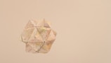 A geometric, paper-crafted object with a complex, polyhedral shape is suspended against a beige background. The object appears to be made from pieces of paper with map-like details, featuring lines and small symbols.
