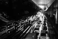 A nighttime urban scene depicts heavy traffic on a wet city road, with bright headlights reflecting off the wet surface. Cars are lined up closely in multiple lanes, and a large overhead structure is visible.