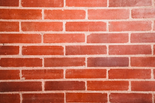 A close-up of a red brick wall with uniform rectangular bricks. The bricks are arranged in a staggered horizontal pattern with light-colored mortar in between.