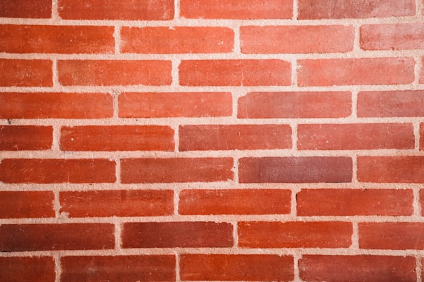 A close-up of a red brick wall with uniform rectangular bricks. The bricks are arranged in a staggered horizontal pattern with light-colored mortar in between.